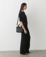 Model wearing a top and pants, holding a bag with a long strap.