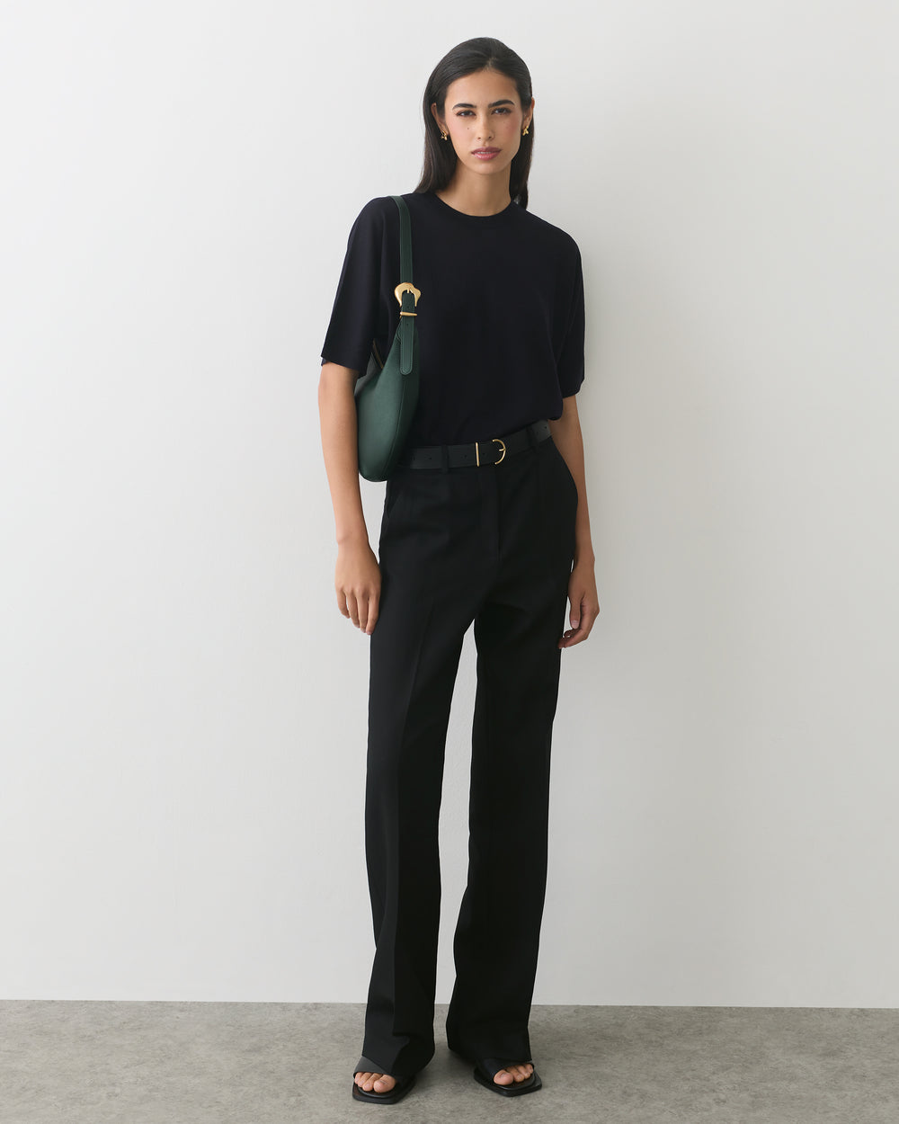 Person standing against a white wall wearing a t-shirt, trousers, and carrying a shoulder bag.