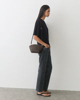Person standing sideways, wearing a T-shirt, jeans, sandals, and carrying a small shoulder bag.