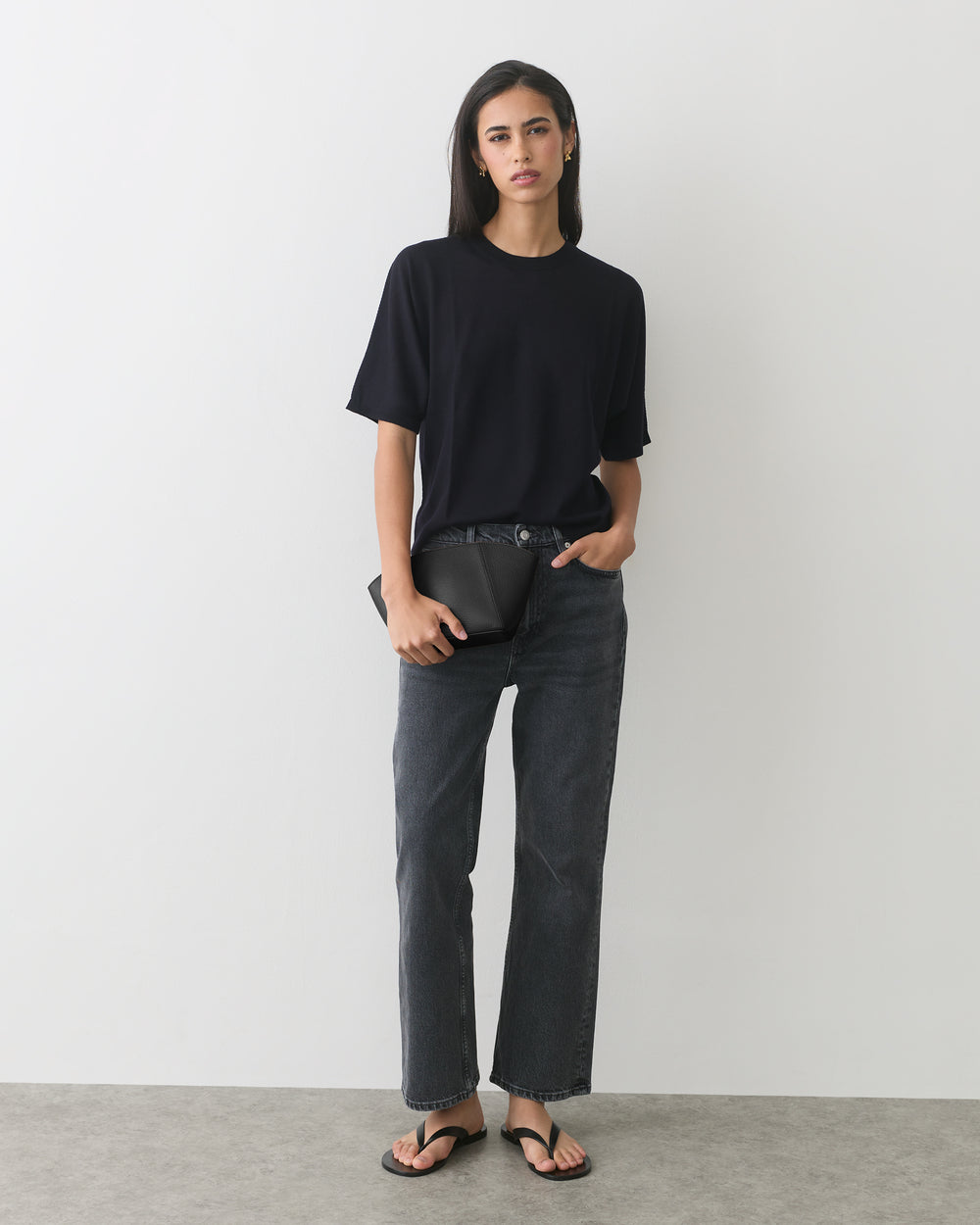 Person standing wearing a T-shirt, jeans, and sandals, holding a clutch.