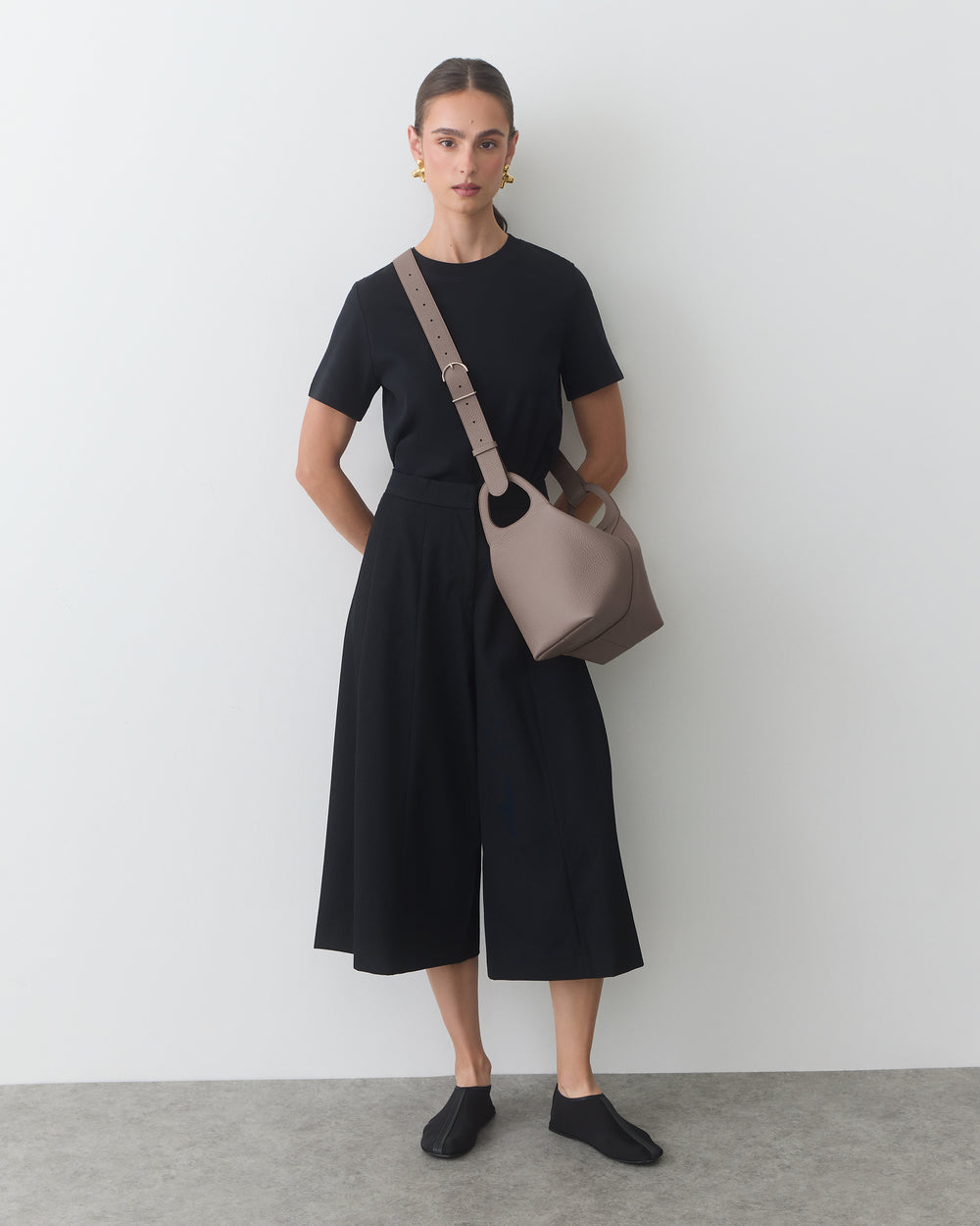 Person wearing a short-sleeve shirt, wide-leg pants, bag and slip-on shoes standing against wall