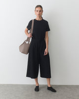 Person standing, holding a travel bag, wearing a T-shirt and wide-leg pants.