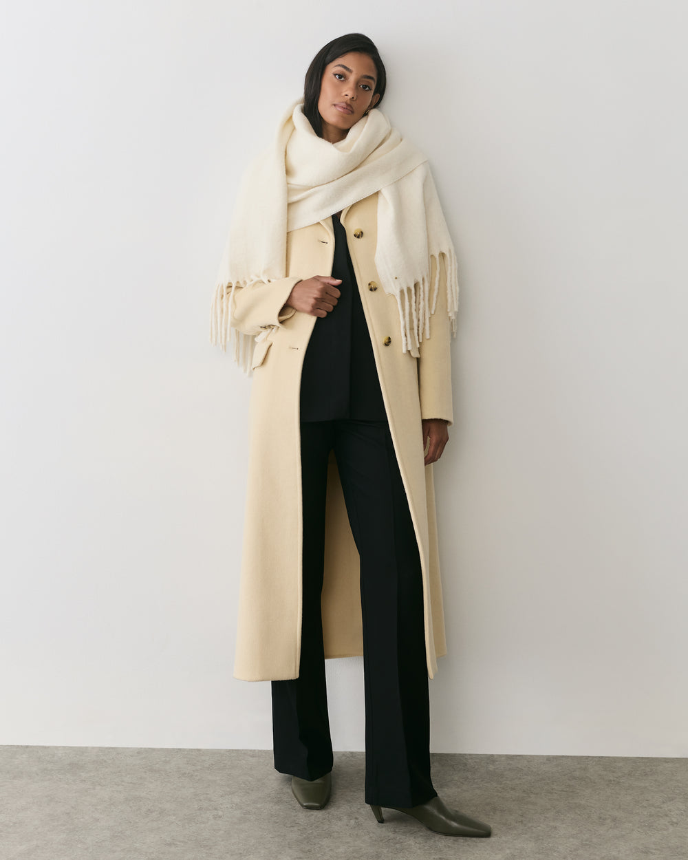 Person wearing long coat, scarf, and trousers stands against plain wall
