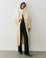 Person wearing long coat, scarf, and trousers stands against plain wall