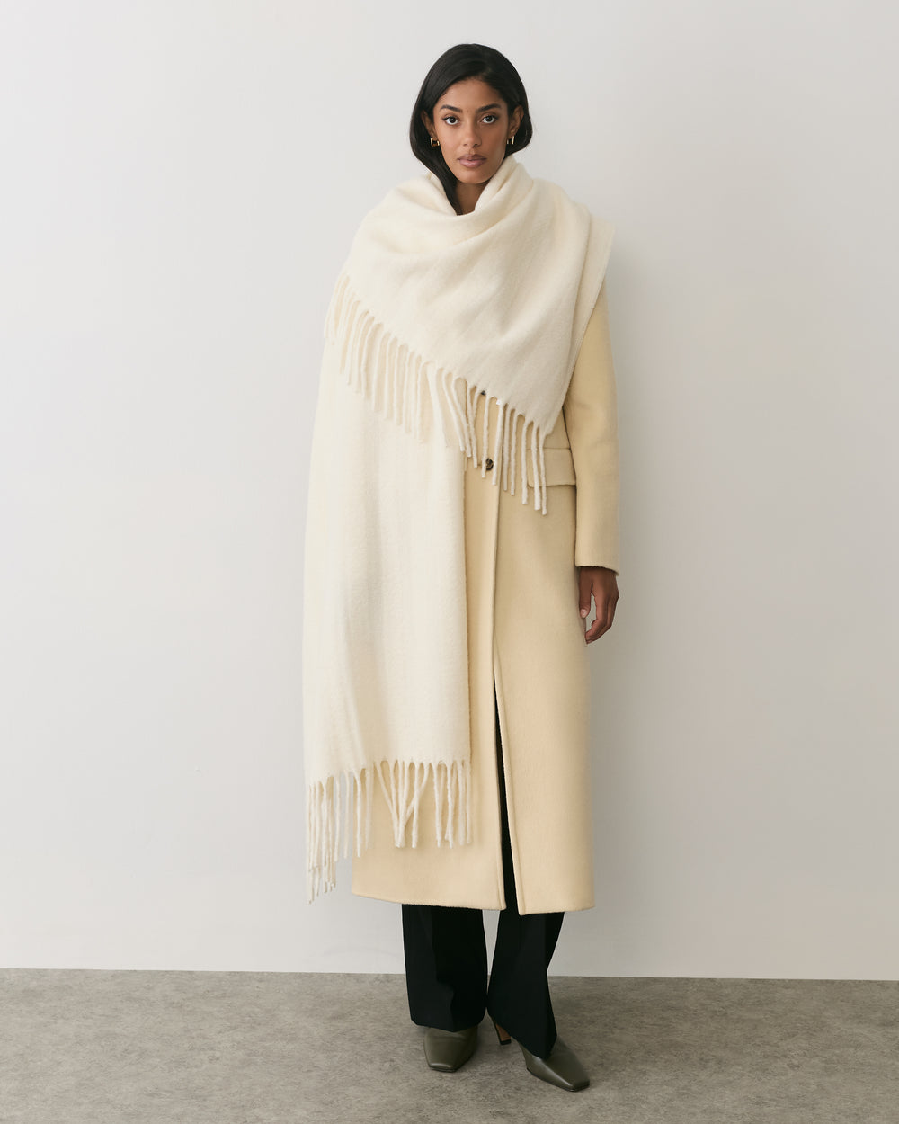 Person wearing long coat and large scarf standing in a plain setting.