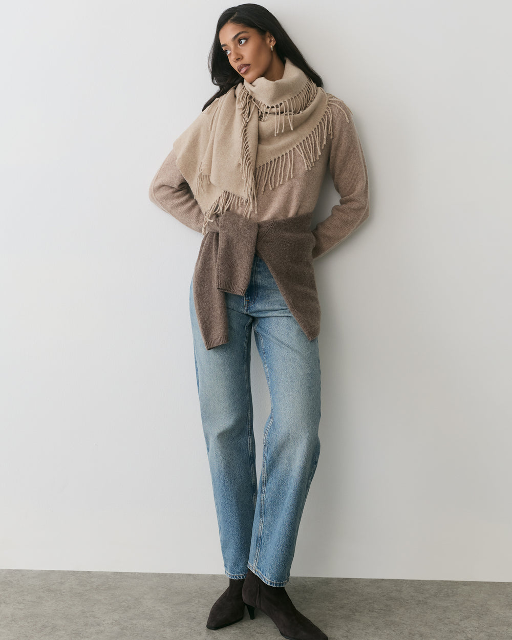 Person wearing a scarf, sweater, and jeans, standing against a plain background