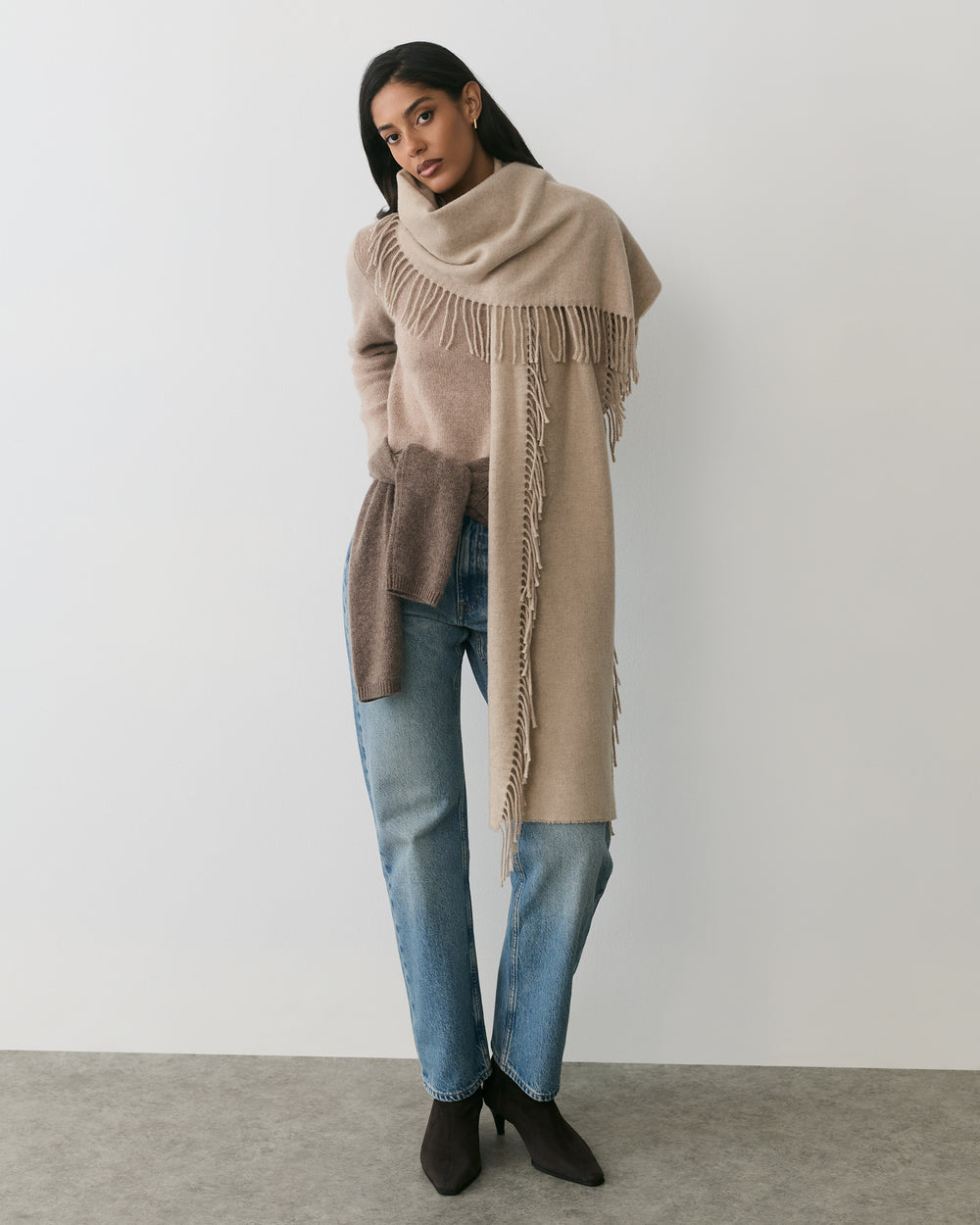 Person wearing a scarf, sweater, jeans, and boots stands against a plain backdrop.