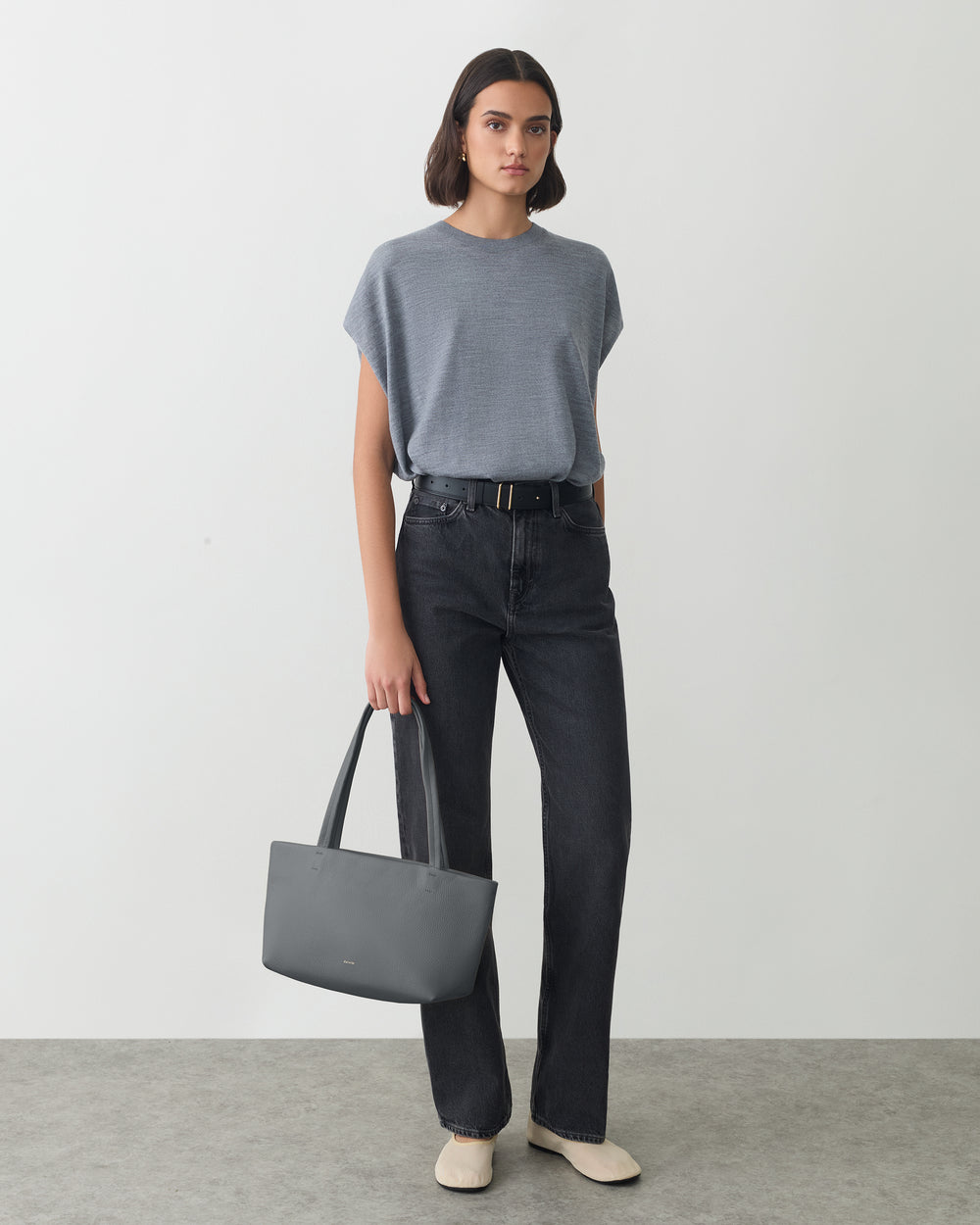 Person stands holding a handbag, wearing a t-shirt and jeans.
