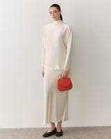 Person in long outfit holding a small purse, standing against a plain backdrop.