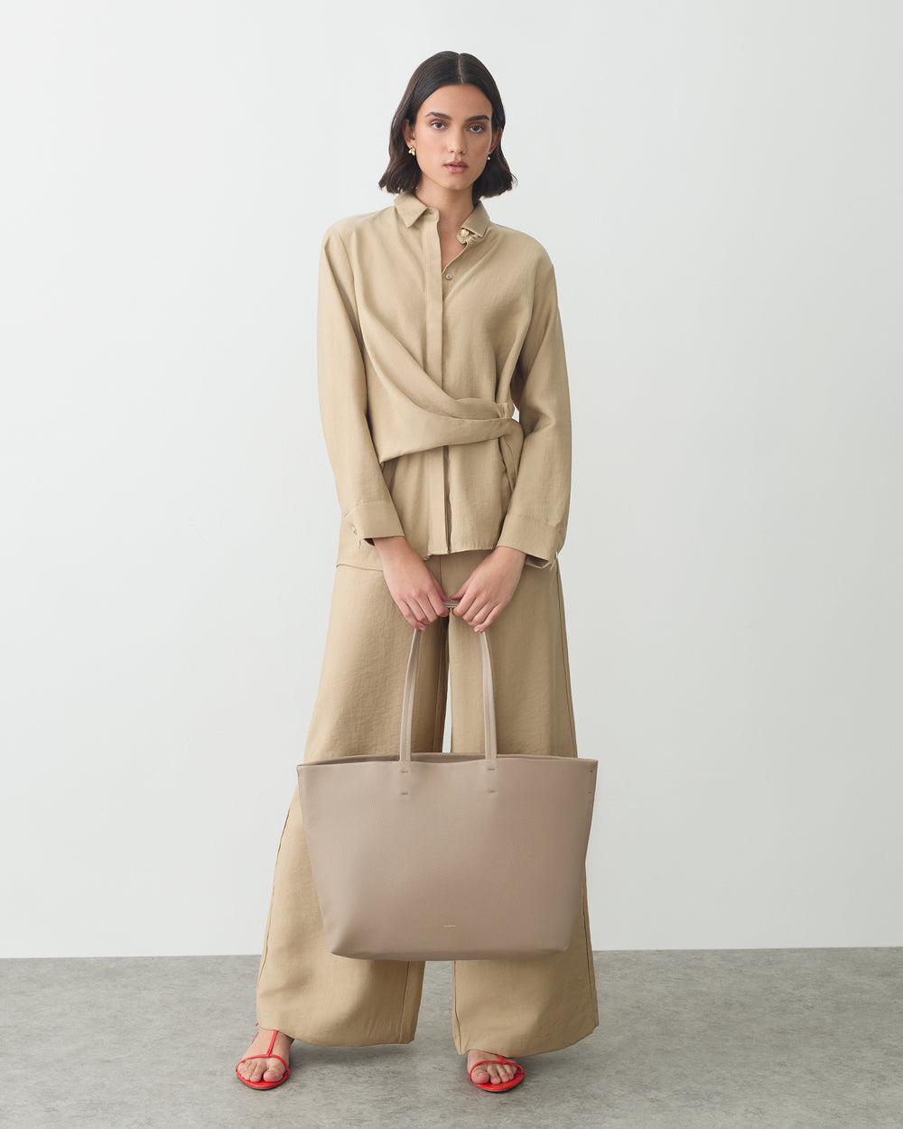 Person standing with a large handbag wearing a belted top and loose pants.