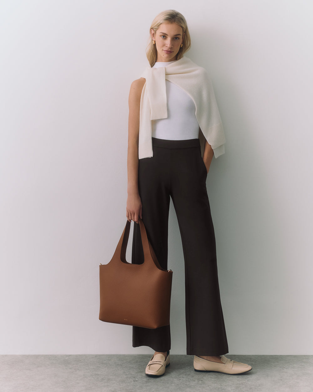 Person stands against a wall, holding a bag, with a sweater draped over shoulders.