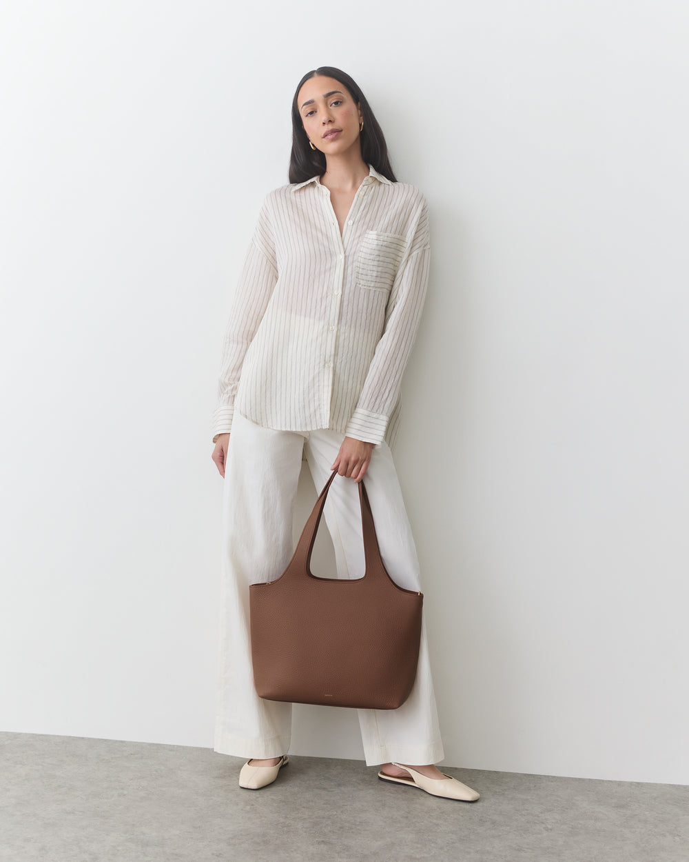 Person in button-up shirt and pants holding a handbag, standing against a plain background.