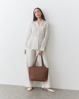 Person in button-up shirt and pants holding a handbag, standing against a plain background.