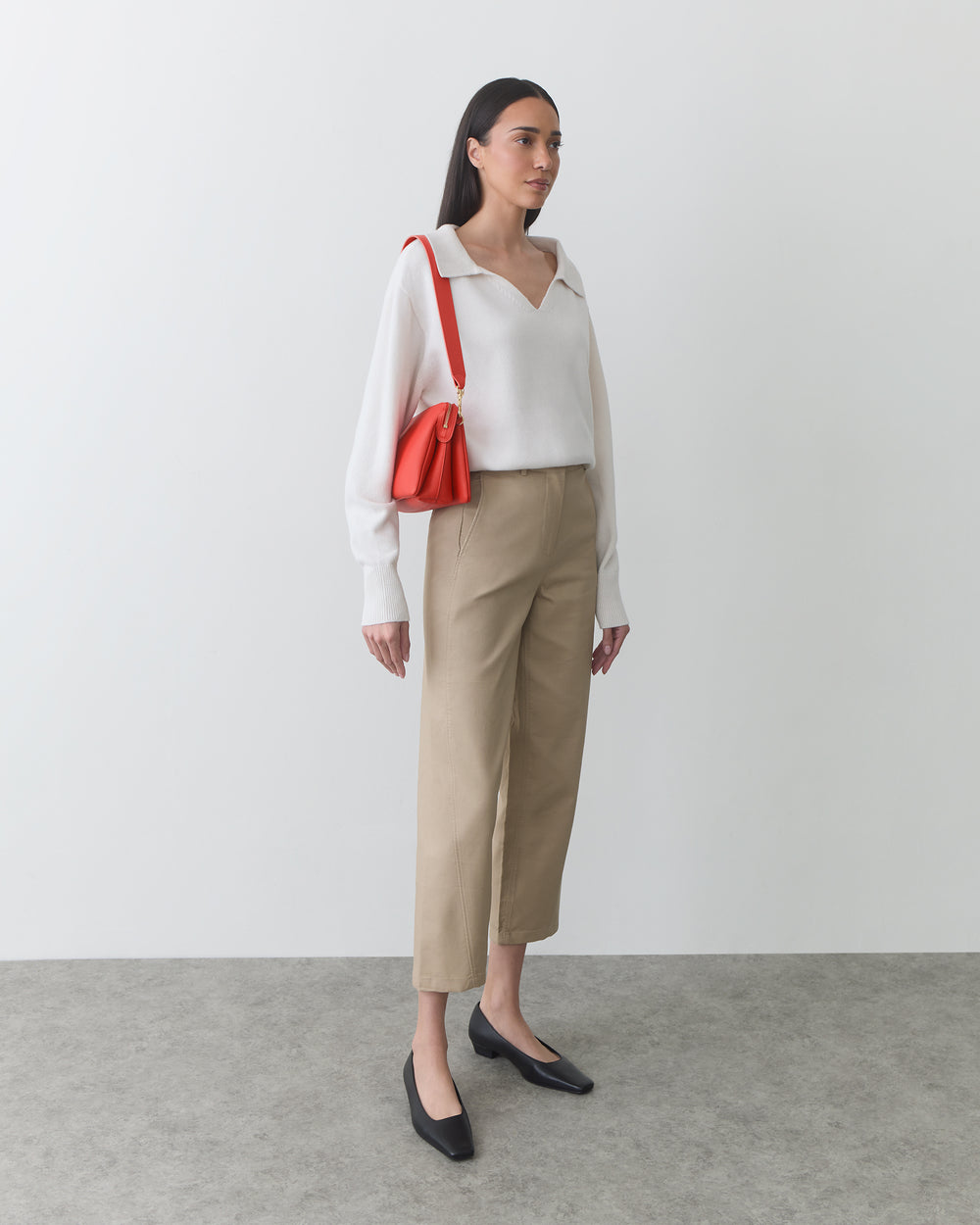 Person wearing a long-sleeve top, pants, holding a shoulder bag, standing indoors.