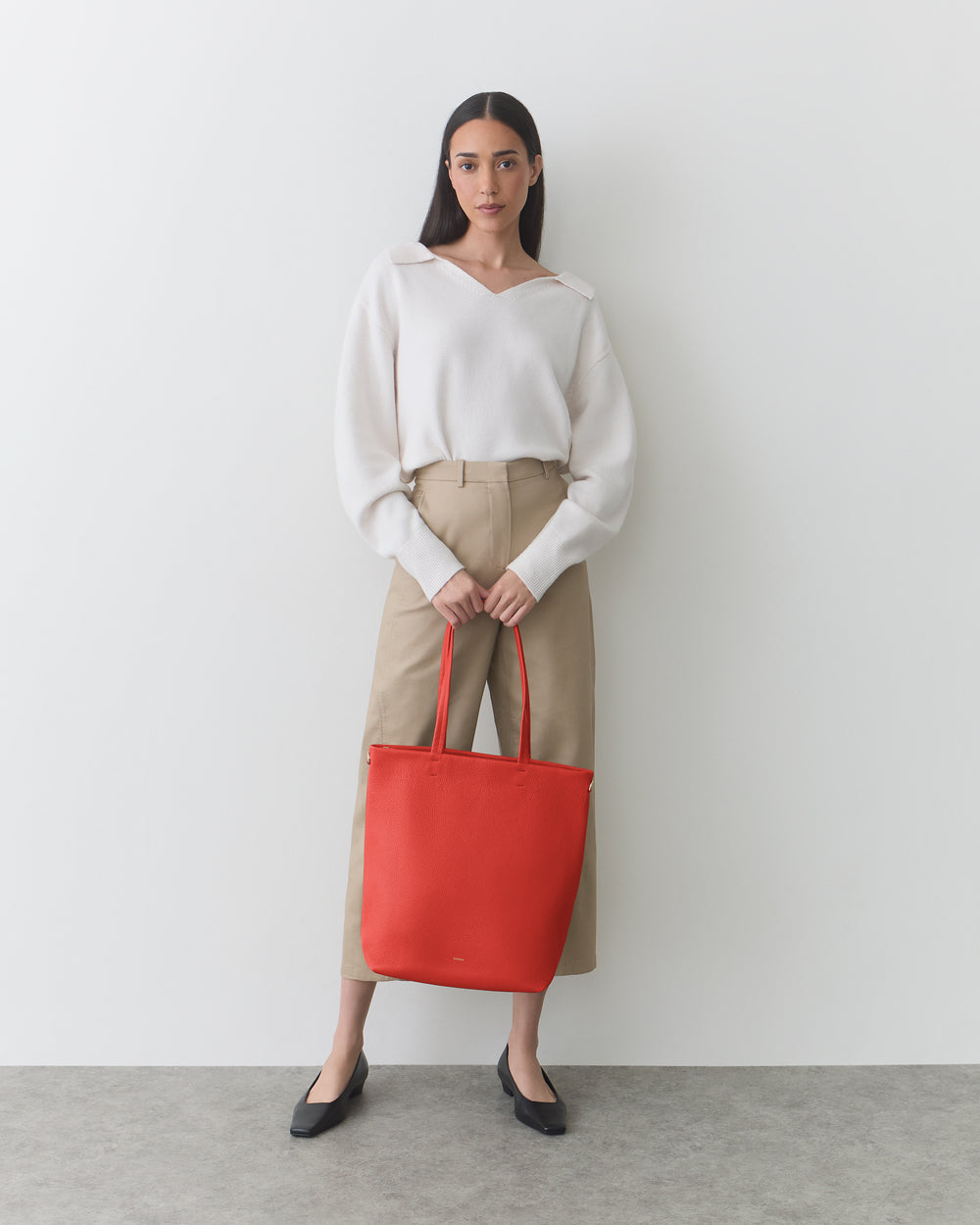 Person standing, holding a large tote bag with both hands, wearing long-sleeve top and pants.