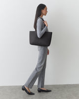 Person standing with a tote bag on shoulder, wearing a long-sleeve top and pants.