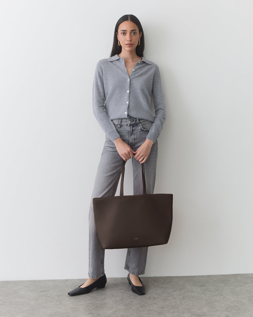 Person standing against a wall holding a large tote bag, wearing a shirt and jeans.