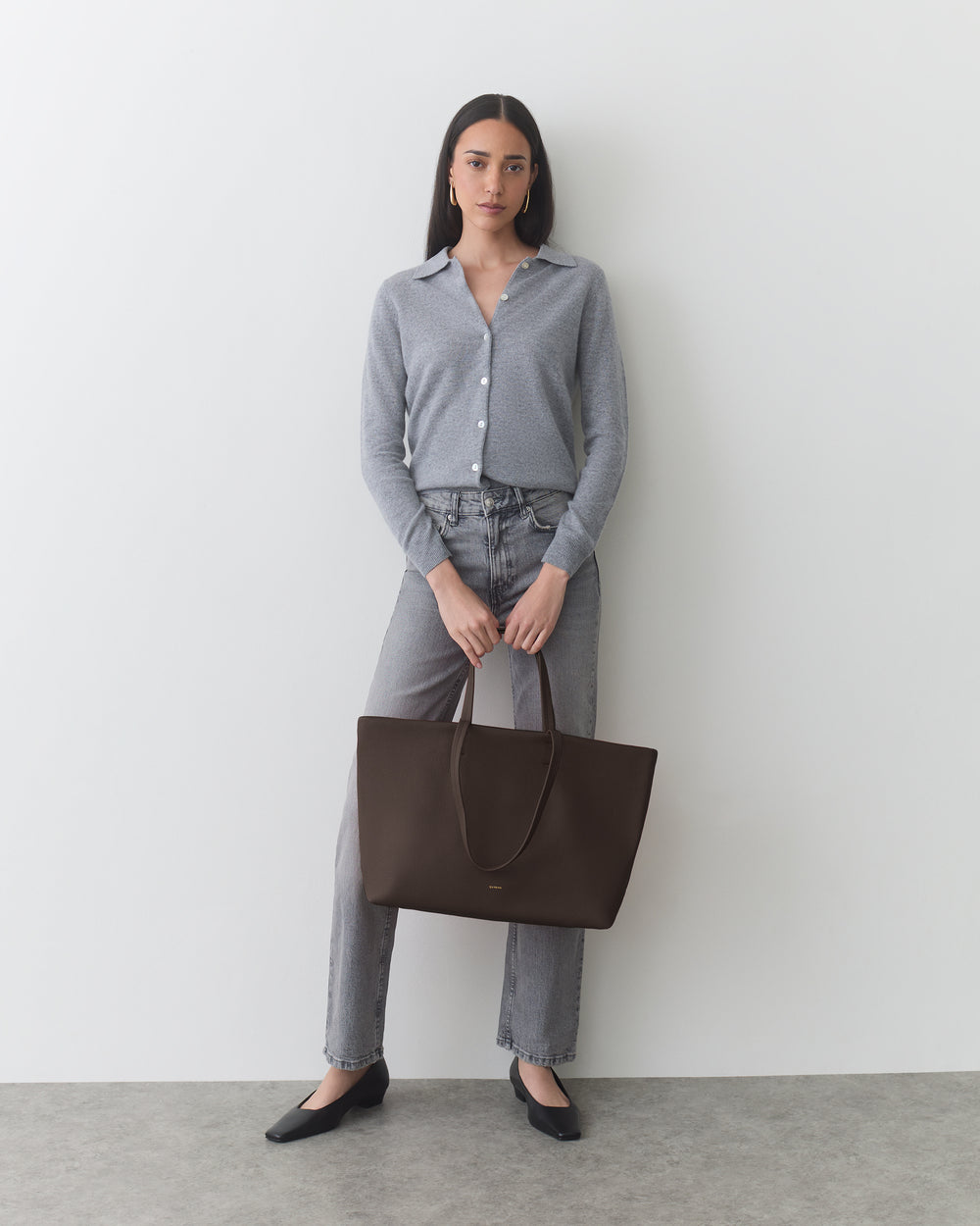 Person standing with a large tote bag, wearing a button-up and pants.