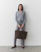 Person standing with a large tote bag, wearing a button-up and pants.