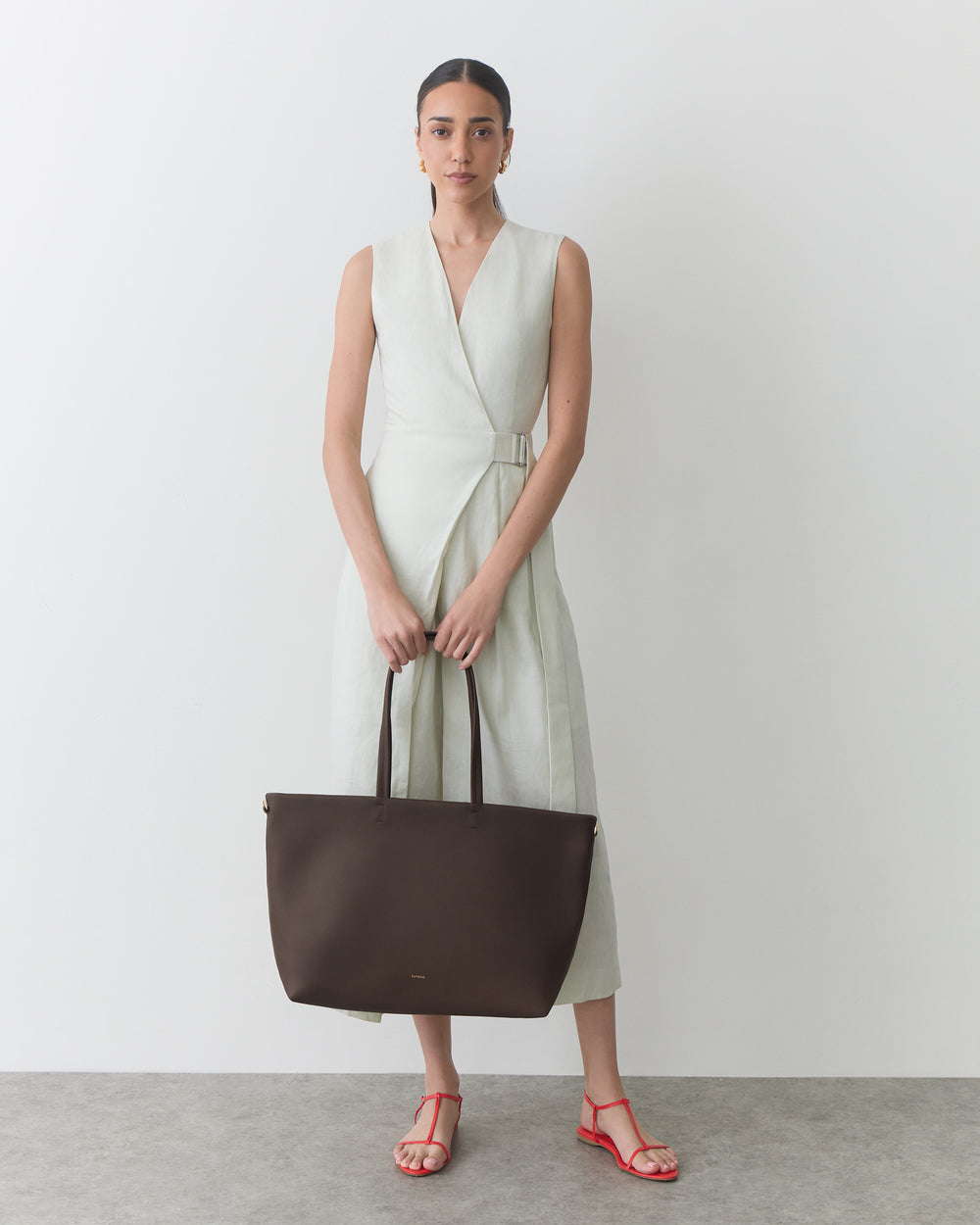 Person in sleeveless dress holding a large handbag, standing on a plain background.