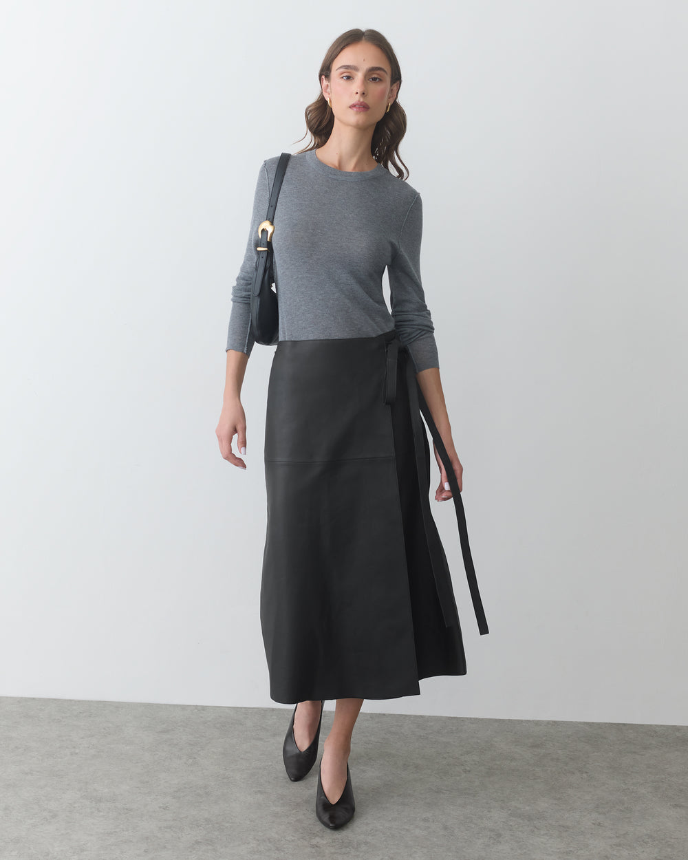 Person in long sleeve shirt and wrap skirt holding a bag, standing indoors.