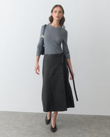Person in long sleeve shirt and wrap skirt holding a bag, standing indoors.