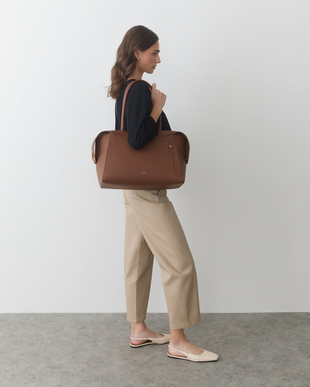 Person standing, holding a large handbag, wearing pants and a long-sleeve top.