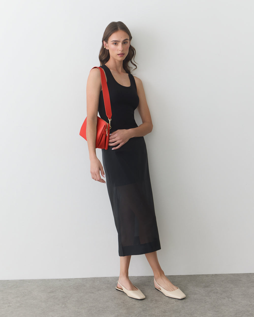 Woman in long dress with shoulder bag and slip-on shoes stands against a wall.