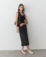 Woman in sleeveless dress with a shoulder bag and flat shoes standing against a wall.