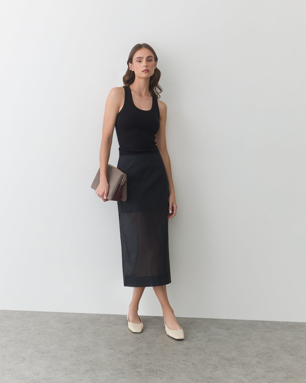 Woman in sleeveless top and skirt holding envelope clutch and standing against blank wall.