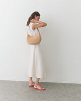 Person in a long dress holding a shoulder bag, standing against a plain backdrop.