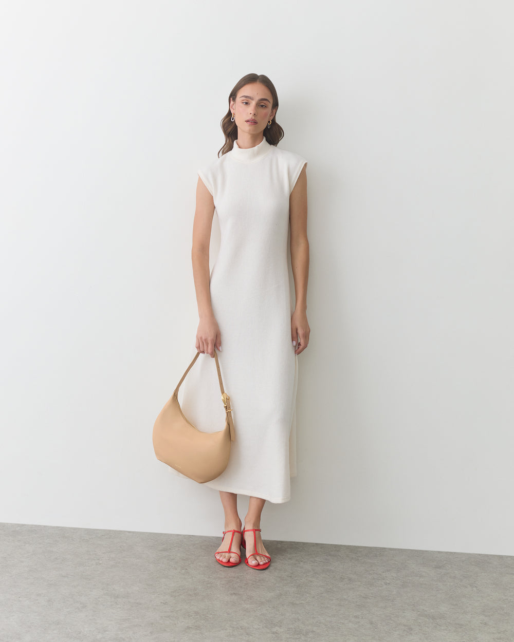 Woman in sleeveless dress holding a shoulder bag, wearing strappy sandals, stands by a wall.