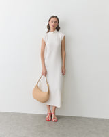 Woman in sleeveless dress holding a shoulder bag, wearing strappy sandals, stands by a wall.