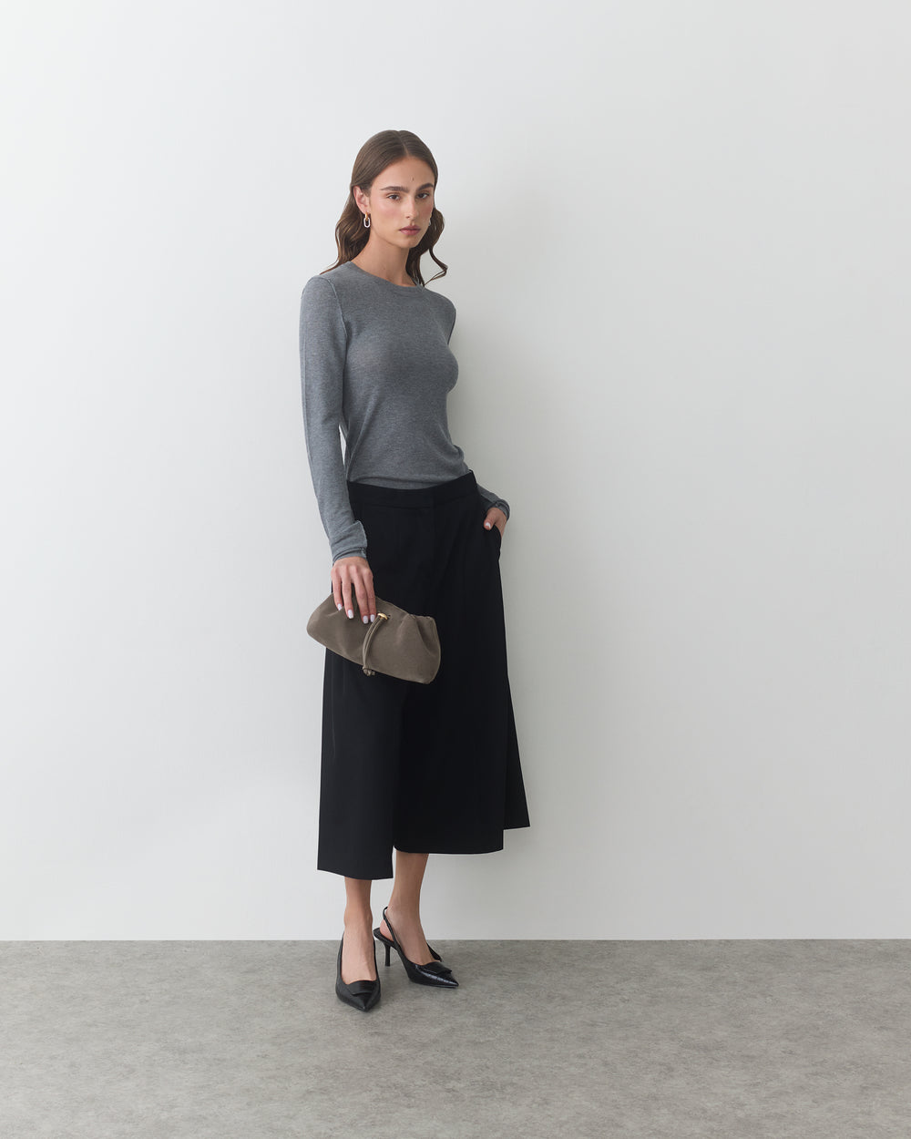 Woman standing in a gray top and black pants holding a clutch bag.