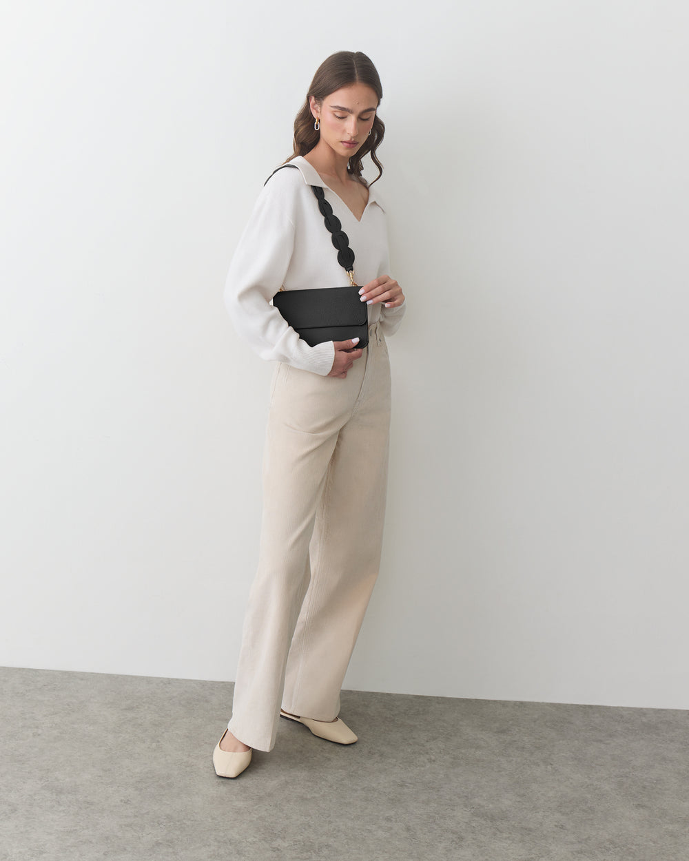 Person in a room holding a purse, wearing a long-sleeve top and wide-leg pants.