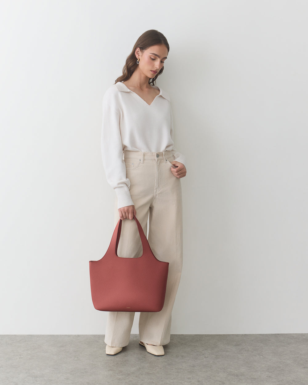 Person holding a tote bag, dressed in a top and pants, standing indoors.
