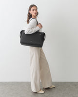 Person standing with a travel bag on shoulder, wearing a long-sleeve top and wide-leg pants.