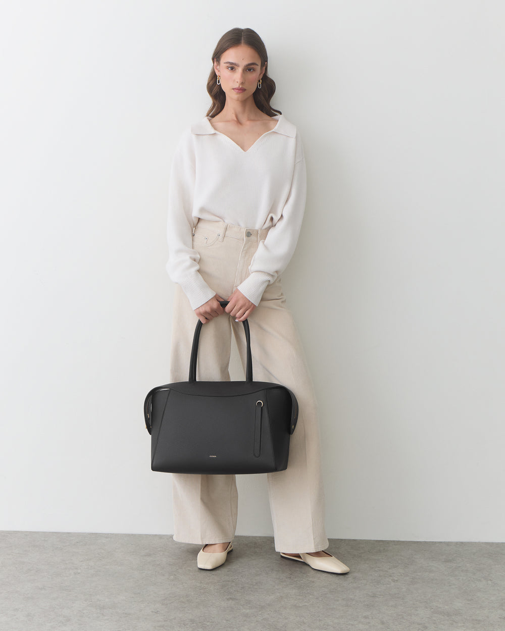 Person standing, holding a handbag, wearing a long-sleeve top and wide-leg trousers.