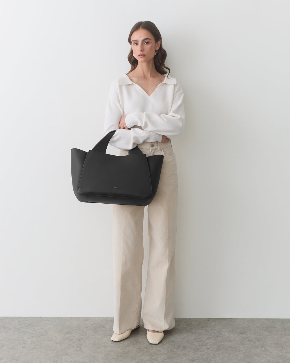 Person holding large tote bag, dressed in long pants and a top, standing against plain backdrop.