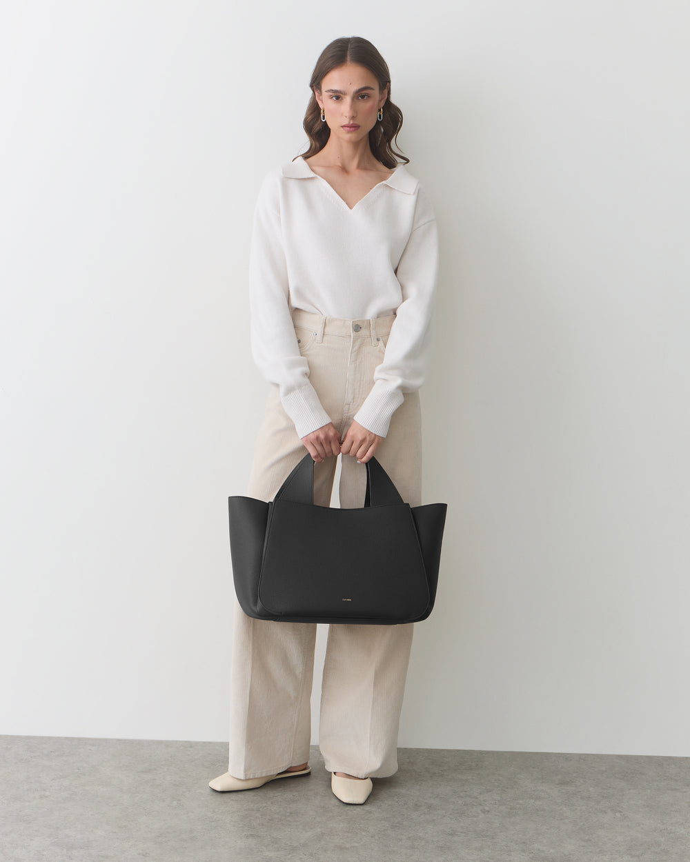 Person wearing a sweater and pants, holding a handbag, standing against a plain wall.