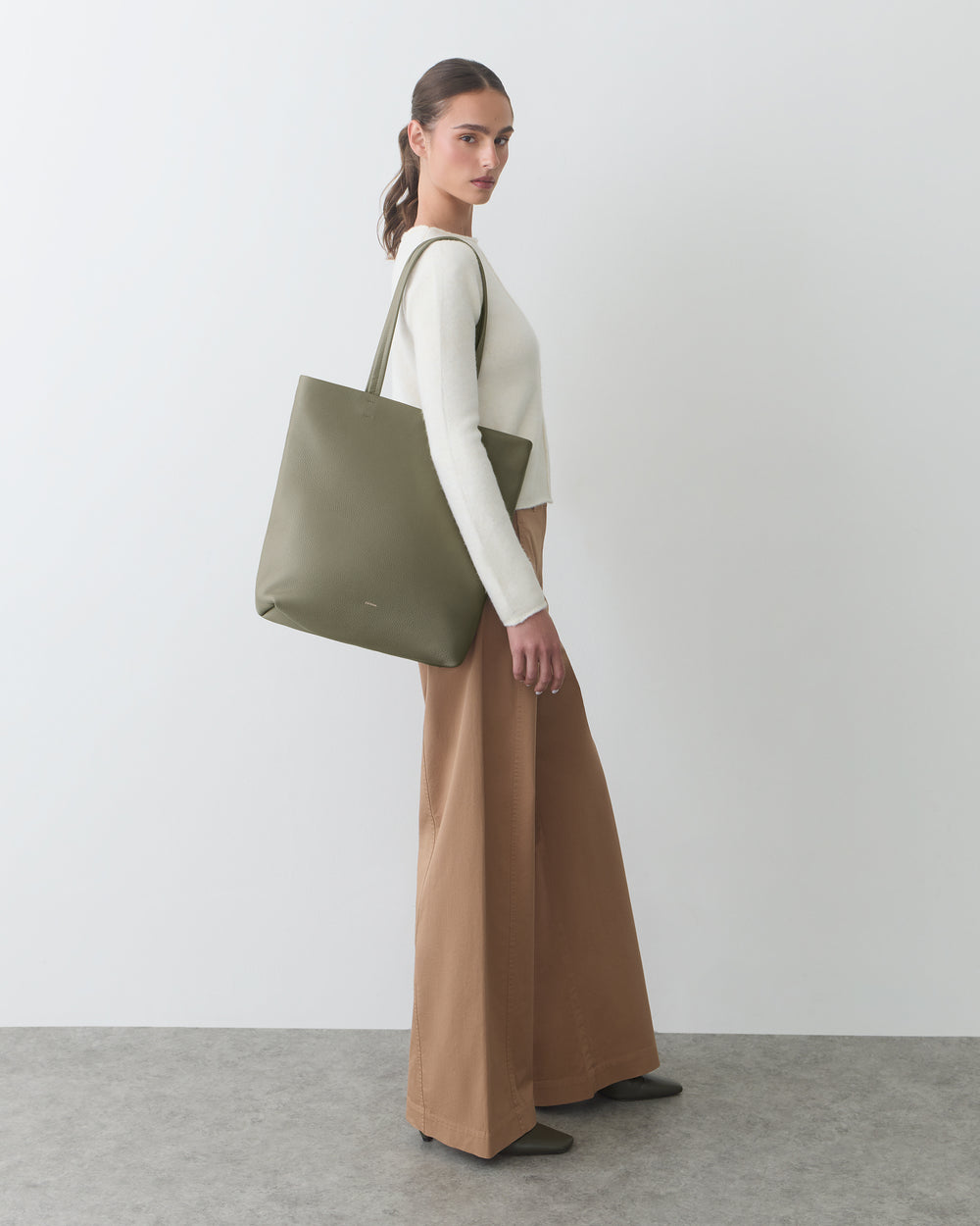 Person with tote bag, wearing long-sleeve top and wide-leg pants, standing sideways.