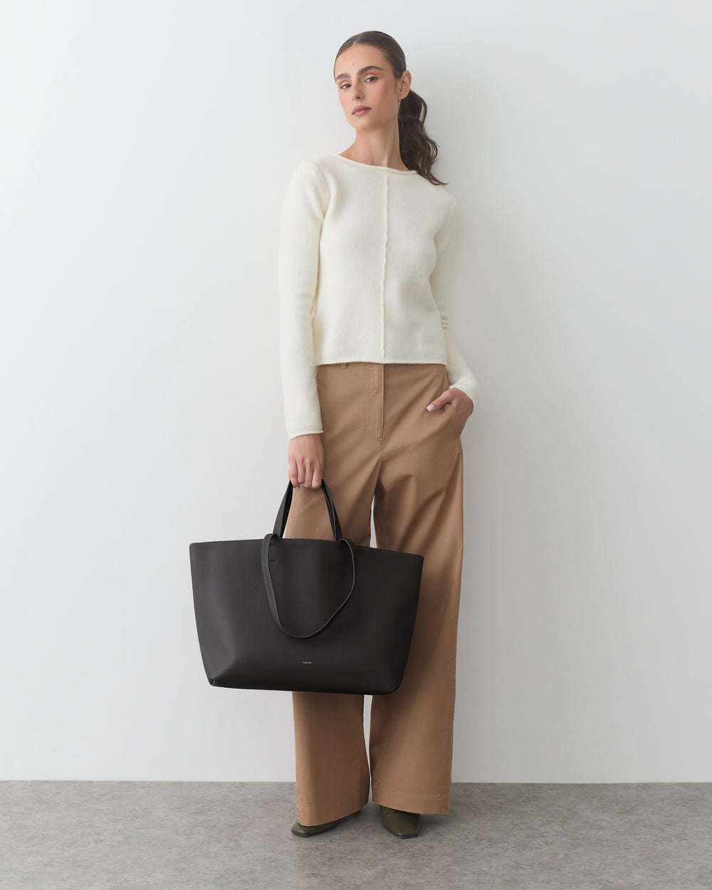 Person in sweater and wide pants holds large handbag, standing against plain background.