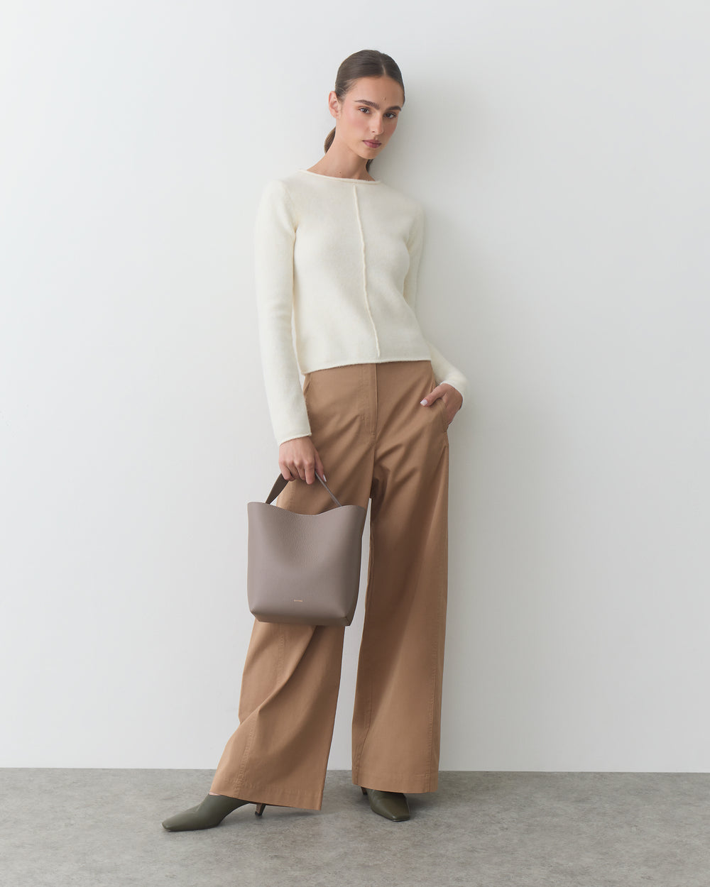 Person posing with wide-leg pants and holding a bag against a plain background.