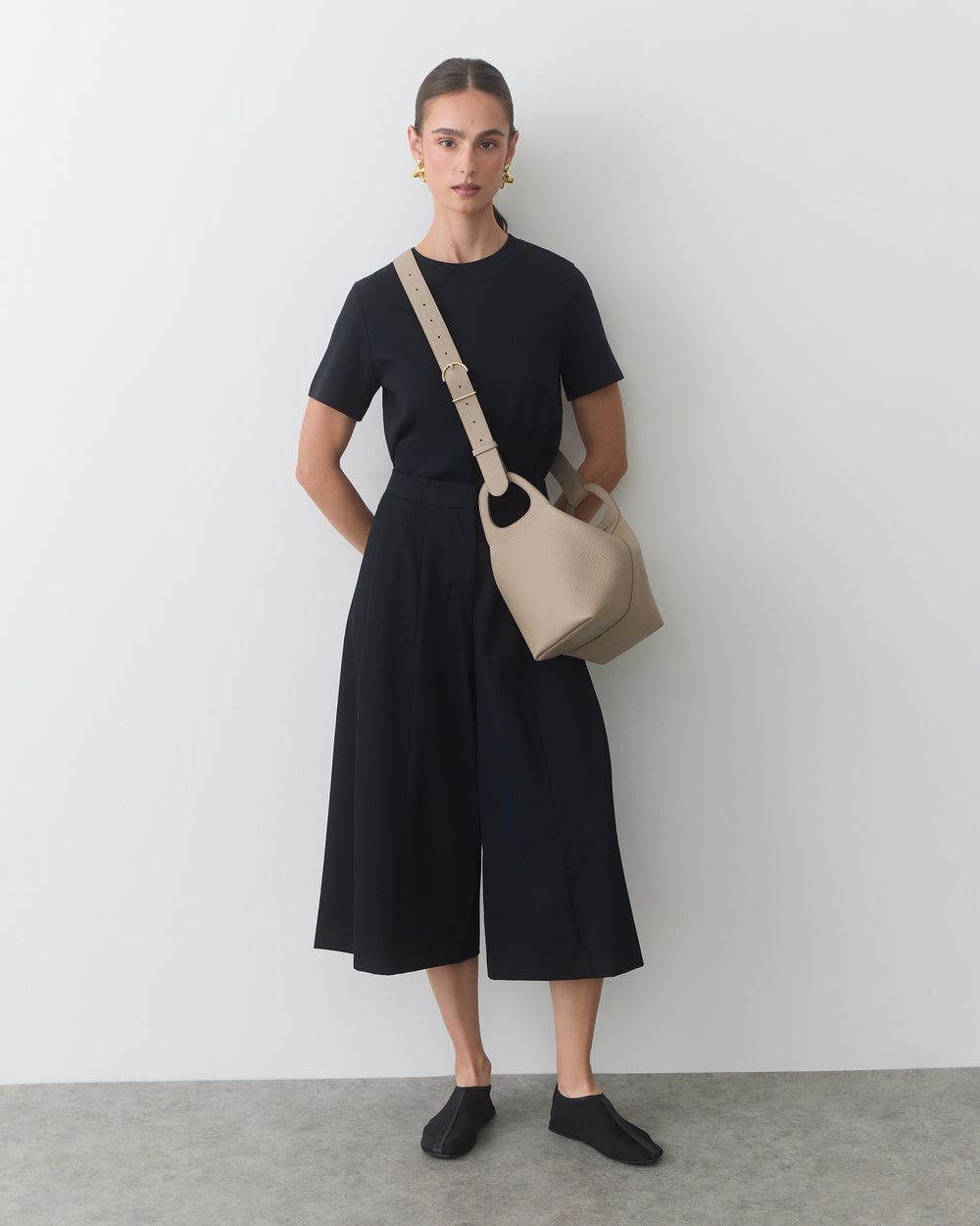 Person in black outfit with crossbody bag stands indoors.