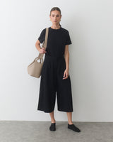 Person standing with a travel bag over shoulder, wearing a short-sleeve shirt and culottes.