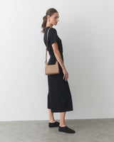 Person standing with a small shoulder bag, wearing a knee-length dress and slip-on shoes.