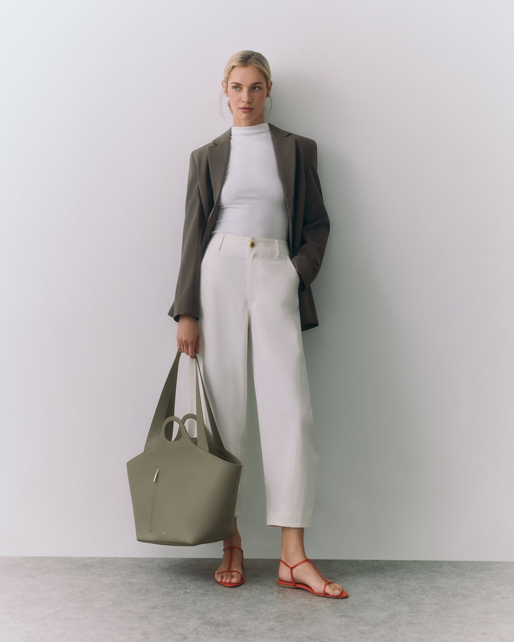 Person in blazer and pants holding a bag, standing against a plain backdrop.