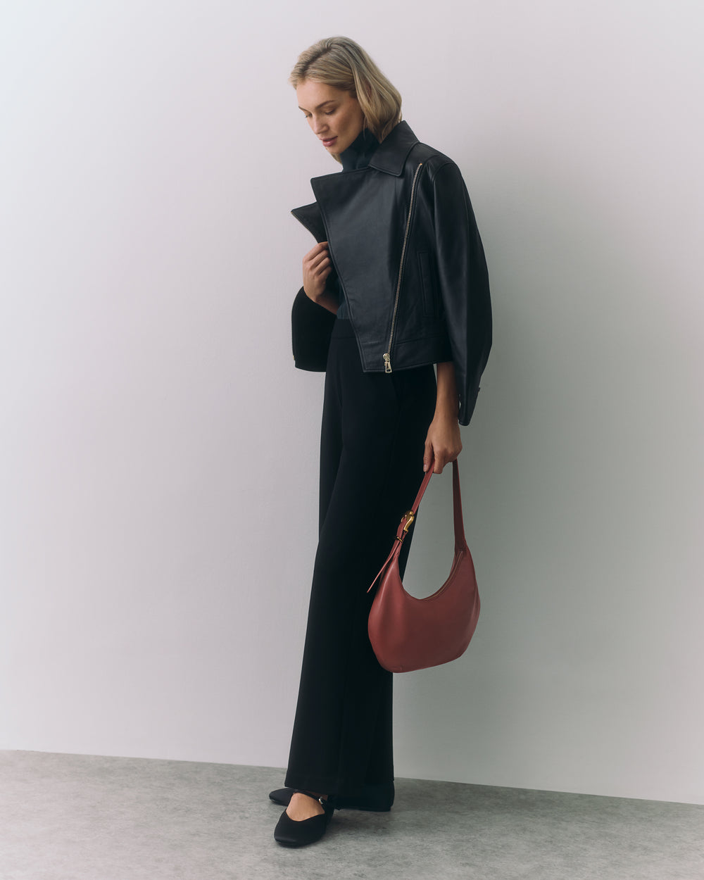 Person wearing jacket and pants holding a handbag standing in a minimalistic setting.