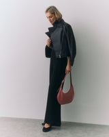 Person wearing jacket and pants holding a handbag standing in a minimalistic setting.