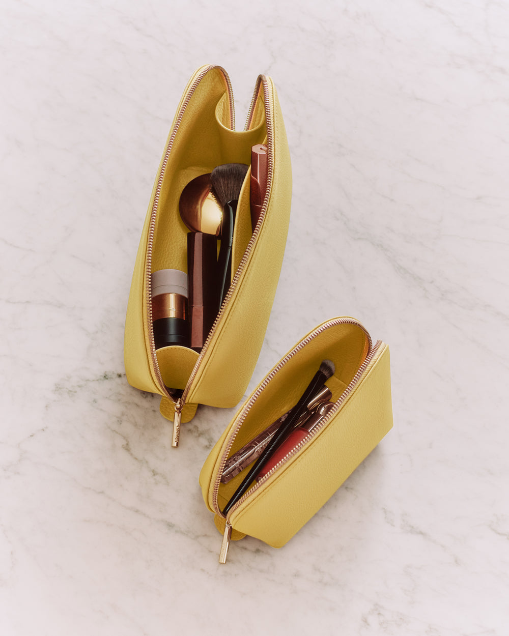 Two open makeup bags with brushes and cosmetics on a marble surface.